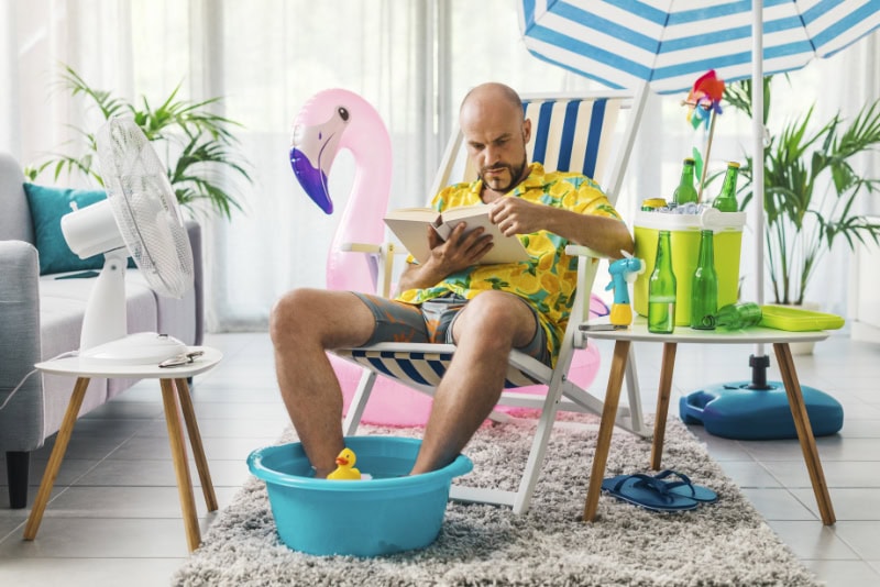 Man spending his summer vacations at home, he is sitting on a deckchair in the living room and reading a book