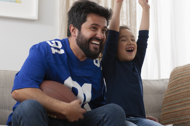 Top 10 Ways to Reduce Your Energy Bill in 2024. Image is a photo of a father and young daughter cheering at a football game on TV.